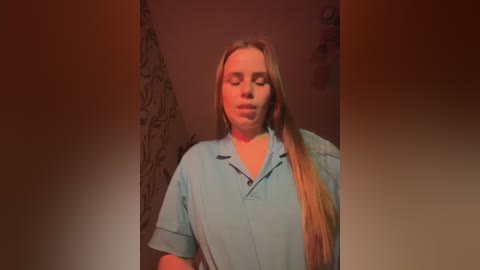 Media: A video of a young woman with long blonde hair, wearing a light blue hospital gown, standing in a dimly lit room with a floral-patterned wall and a blurred background.