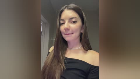 Media: Video of a smiling Caucasian woman with long, straight brown hair, wearing a black off-shoulder top, in a dimly lit hallway.
