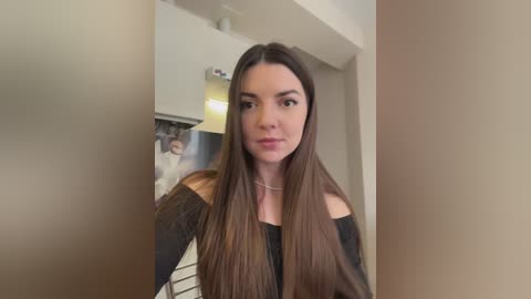 Media: A video of a fair-skinned woman with long, straight brown hair, wearing a black off-shoulder top, standing in a modern kitchen with white cabinets and a blurred background.