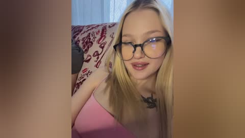 Media: Video of a young woman with long blonde hair, fair skin, and glasses, wearing a pink top, sitting on a patterned chair, looking directly at the camera.