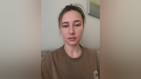 Video of a young woman with light skin and brown hair tied back, wearing a plain brown T-shirt. She sits indoors against a beige wall, with a blurred painting in the background.