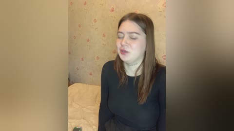 Media: A video of a young Caucasian woman with long brown hair, fair skin, and a small build, wearing a black top, sitting on a beige cushion in a room with floral-patterned wallpaper.