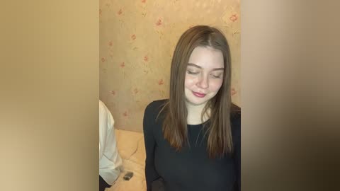 Media: A video of a young woman with light skin and straight, shoulder-length brown hair, wearing a black top, standing in a small, dimly lit room with beige floral wallpaper and a bed with white sheets.