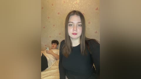 Media: Video of a young woman with straight brown hair and fair skin, wearing a black long-sleeved top, standing in a beige room with floral wallpaper. A child in a yellow blanket is lying on a bed in the background.
