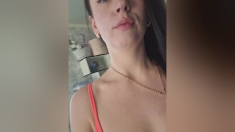 Video of a young Caucasian woman with light skin, long dark hair, wearing a red spaghetti strap top, and a gold necklace. Background shows a blurred room with shelves and decorative items.