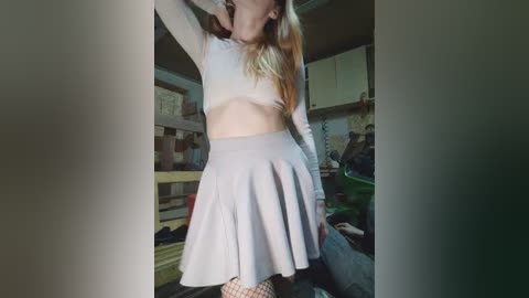 Video of a slim, fair-skinned woman with long blonde hair, wearing a white crop top and a short, pleated skirt. She is in a cluttered, dimly-lit room with shelves and clothes visible in the background.