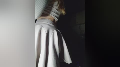 Video of a woman from the waist down, wearing a white pleated skirt and grey knit top, standing in dimly lit room with shadowed background.