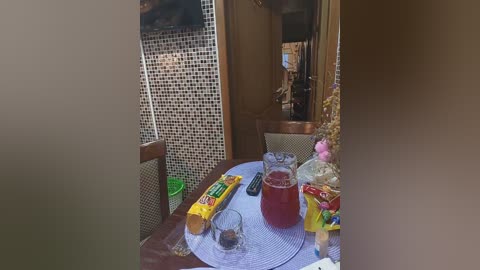 Media: Video of a small, cluttered dining area with a round wooden table. The table is covered with a white placemat, featuring a glass of red wine, a clear glass, a yellow bag, and a green bowl. The background includes a mosaic-tiled wall and wooden furniture.