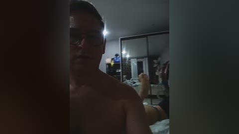 Media: A dimly lit video shows a shirtless man with short dark hair, wearing glasses, standing in a cluttered bedroom with a mirror reflecting his partially obscured reflection.