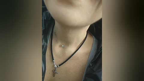 Video of a person's neck and chest, showing a black necklace with a crucifix pendant, and a belly button piercing. The background is blurred and dark.