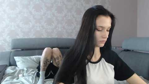 Media: A video of a young woman with long black hair, wearing a white and black t-shirt, lying on a gray couch with floral wallpaper in the background.