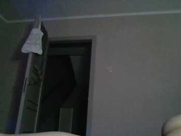 Video of a dimly lit, sparsely furnished room with a beige wall, a metal ladder leaning against it, and a partially open door leading to a darker space.