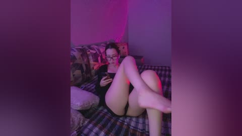 Media: Video of a woman with glasses and fair skin, lying on a checkered bed in a dimly lit room with purple lighting, wearing black lingerie.