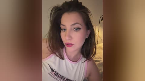 Video of a young woman with fair skin, dark hair, and striking blue eyes, wearing a white tank top with a pink trim, standing indoors against a blurred background.