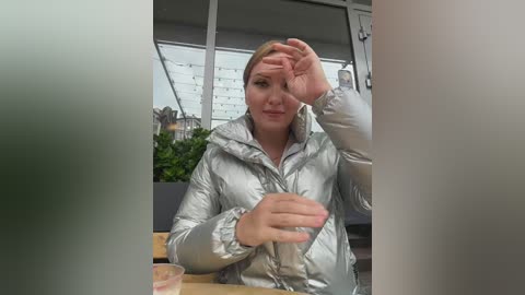 Video of a blonde woman in a silver puffer jacket, shielding her eyes with her right hand, sitting at a wooden table with a glass of red liquid in a cafe.