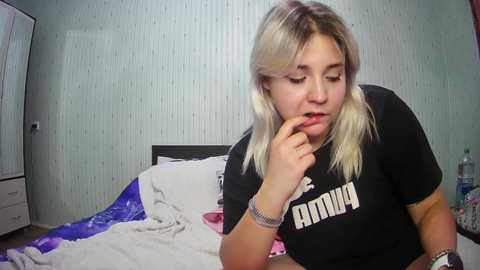 Media: Video of a blonde woman with fair skin, wearing a black t-shirt, lying on a bed, looking contemplative. Background includes a white dresser and a partially visible mirror.