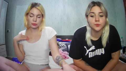 Media: Video of two young women with light skin, one blonde and one brunette, kissing on a bed. They wear casual shirts and shorts. Background shows a messy room with a Puma logo.