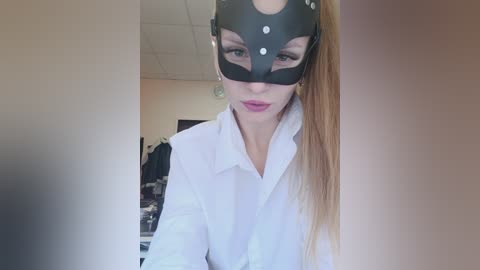 Video of a light-skinned woman with long blonde hair, wearing a black leather mask with white studs, and a white shirt, indoors with a blurred background.