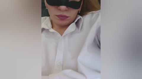 A close-up video of a woman wearing a black blindfold, white shirt, and lipstick, partially obscured by a blurred white object, creating a mysterious, intimate atmosphere.