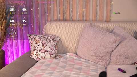Video of a cozy living room with a beige sofa, light pink and white geometric-patterned blanket, and a pillow featuring a cartoon rabbit design. Background includes a curtain with a subtle pink and beige pattern, and a decorative plant.