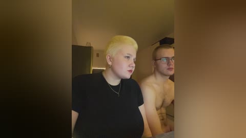 Video of two shirtless men with short hair and glasses standing in a dimly lit room, one wearing a black T-shirt, the other bare-chested.