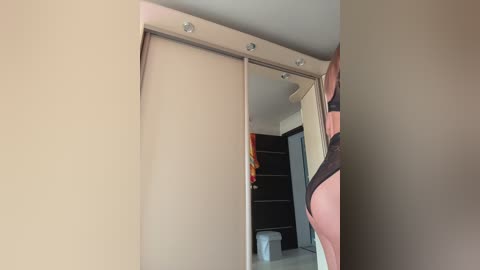 Video of a modern bedroom with beige walls, a sliding wardrobe, and a partially visible person wearing black lingerie, standing in front of the wardrobe, viewed from the side.