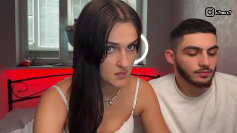 Video of a young woman with long dark hair and a serious expression, standing next to a man with short dark hair and a beard, in a modern bedroom with red lighting.