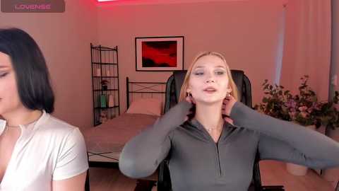 Video of a blonde woman in a grey hoodie, sitting on a black office chair, with another woman in a white top in the background, in a cozy, dimly lit room with a bed, plants, and framed art.