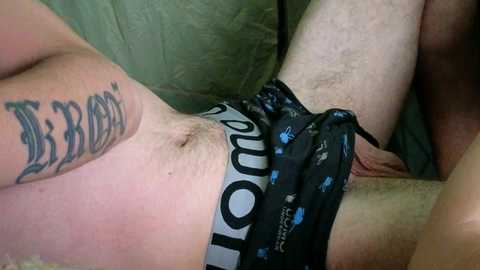 Media: Video of a person with light skin and a tattoo on their upper arm, wearing black and blue boxer shorts, lying on a green couch.