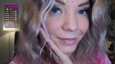 Media: Video of a smiling woman with wavy, light blonde hair, blue eyes, and light skin, wearing a pink top. Background includes a digital display with text and icons.