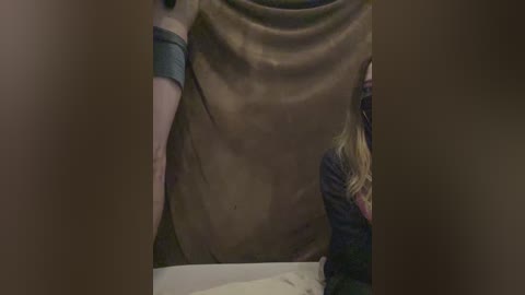 Video of a person wearing a brown, torn shirt with a grey sleeve, and a blonde woman in the background, partially obscured by a wooden frame.