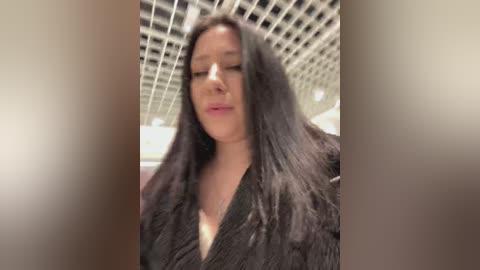 A video of a young woman with long, dark hair, wearing a black, textured cardigan, standing in an office with a grid-patterned ceiling. She has a neutral expression and is looking slightly downward.