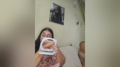 A video of a young woman with long dark hair, wearing a floral dress, holding a white mirror against her breast, revealing her nipple. The background features a framed black-and-white photo and a medical device.