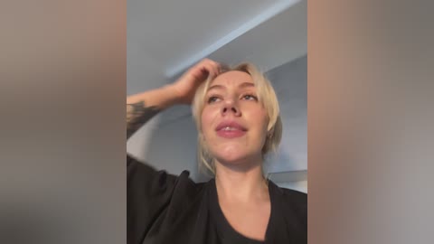 Media: A video of a blonde woman with fair skin, wearing a black top, adjusting her hair with her right hand. The background features a blurred, light-colored room with minimal decor.