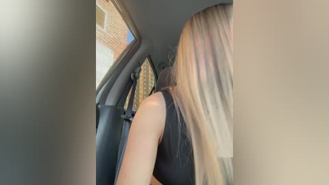Media: Video of a blonde woman with long, straight hair in a black sleeveless top, leaning forward in a car with beige upholstery, sunlight streaming through the window.