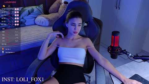 Media: Video of a young woman with pale skin and dark hair, wearing a white strapless crop top and high-waisted black shorts, sitting in a gaming chair with a plush bear and a red webcam in the background.