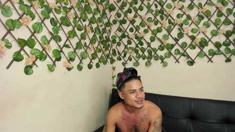 Video of a shirtless, tattooed, muscular Asian man with short black hair and a black headband, leaning back on a black leather couch. He's smiling. The background features a lattice wall adorned with green vines and pink flowers.