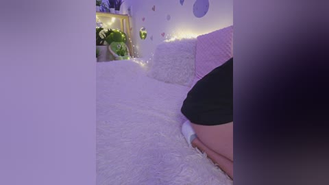 A video of a cozy bedroom with a fluffy white rug, pink and purple bedding, and fairy lights, featuring a partially visible person's leg in black shorts.