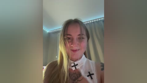 Media: Video of a young, light-skinned woman with long blonde hair and a white sleeveless top, smiling, in a dimly lit room with green curtains.