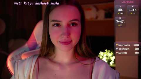 Media: Video of a young Caucasian woman with straight brown hair and fair skin, wearing a white top, sitting in a dimly-lit room with blurred background.