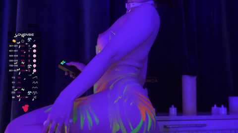 Video of a nude woman with light skin and long dark hair, wearing a multicolored, flowing dress, sitting on a bed with purple lighting. The background includes candles and a TV displaying a virtual reality menu.