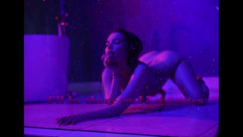 Video of a nude, fair-skinned woman with shoulder-length brown hair, posing on all fours, lit by purple light. She has medium-sized breasts and a slim physique. The background is dimly lit, with a cylindrical object on the left.