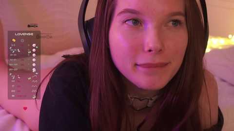Video of a young woman with fair skin, brown hair, wearing headphones, a black top, and a heart necklace, smiling, with a blurred background.