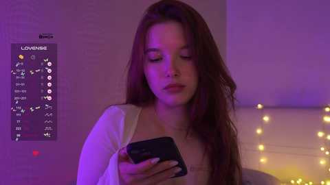 Video of a young woman with long brown hair, wearing a white cardigan, using a smartphone, in a dimly lit room with purple and yellow lights, and a digital clock displaying \"LOVE\" on the wall.