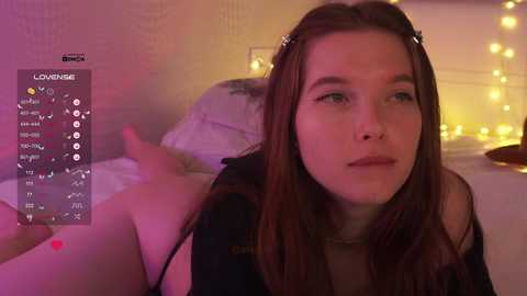 Video of a fair-skinned, young woman with long, auburn hair, lying on a bed with white sheets. She wears a black top, with a soft, ambient lighting creating a cozy, intimate atmosphere.