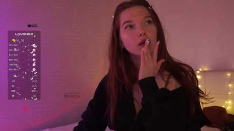 A video of a young woman with long brown hair, wearing a black sweater, in a dimly lit room with a glowing wall and a cozy bed.