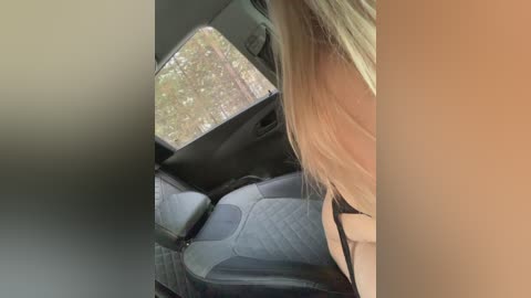 Video of a woman with long blonde hair, seated in a car with gray upholstery. The background shows a blurred forest view through a window.
