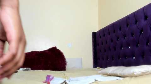 Video of a plush, tufted purple headboard in a minimalist bedroom. A naked person with a light skin tone is seen from the waist down, moving towards the bed. The bed has a beige sheet and a maroon pillow.