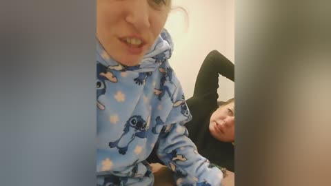 A video of a woman in a blue, patterned pajama top, smiling, with a man partially visible in the background.