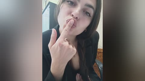 Media: Video of a young woman with fair skin, long brown hair, and large brown eyes, in a black outfit, covering her mouth with her finger. Background shows part of a room with a radiator and a wooden table.